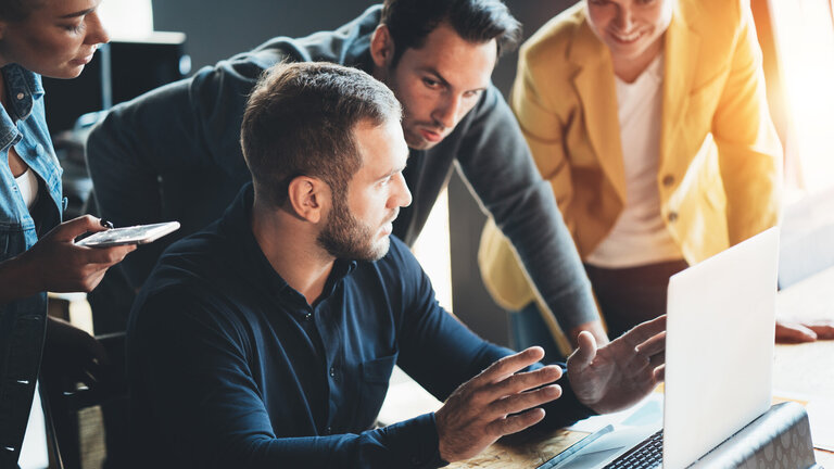 Das Bild zeigt eine Gruppe von vier Personen, die an einem Laptop arbeiten und miteinander interagieren. Ein Mann sitzt im Vordergrund und spricht gestikulierend, während die anderen drei Personen aufmerksam zuhören und sich auf den Bildschirm konzentrieren. Eine Frau hält ein Smartphone in der Hand, vermutlich für Notizen oder zusätzliche Recherchen. Die Szene vermittelt eine dynamische Teamarbeit in einer modernen, professionellen Umgebung.