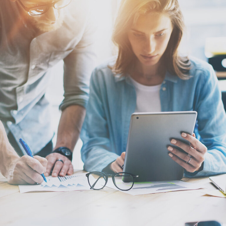 Bürosituation, eine Frau konzentriert sich auf ein Tablet, während ein Mann auf ein Dokument mit Diagrammen zeigt, mit einem Laptop und einer Kaffeetasse im Vordergrund.