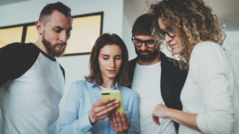 Eine Gruppe von vier Personen steht zusammen und betrachtet konzentriert ein Smartphone.