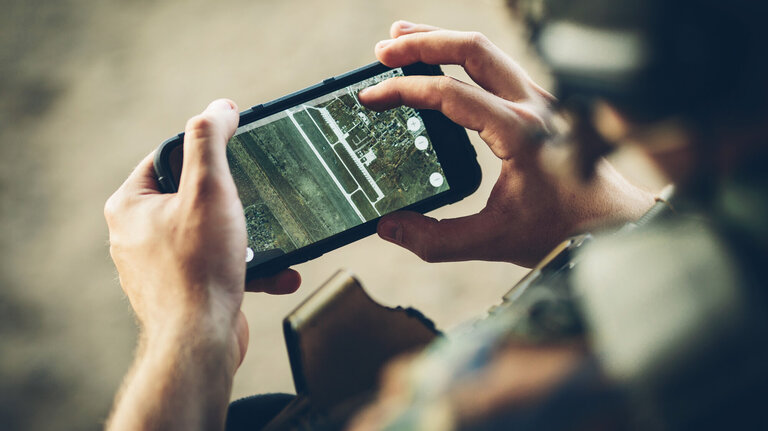 Eine Person betrachtet eine Satellitenkarte auf einem Smartphone, möglicherweise in einer taktischen oder militärischen Umgebung, basierend auf der Kleidung und dem Hintergrund.