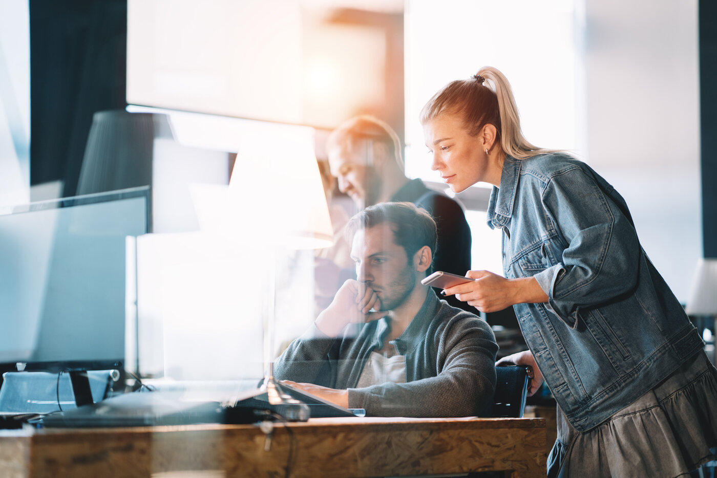 Eine Frau in Jeansjacke zeigt einem Kollegen am Computer etwas auf ihrem Smartphone, während weitere Personen im Hintergrund arbeiten.