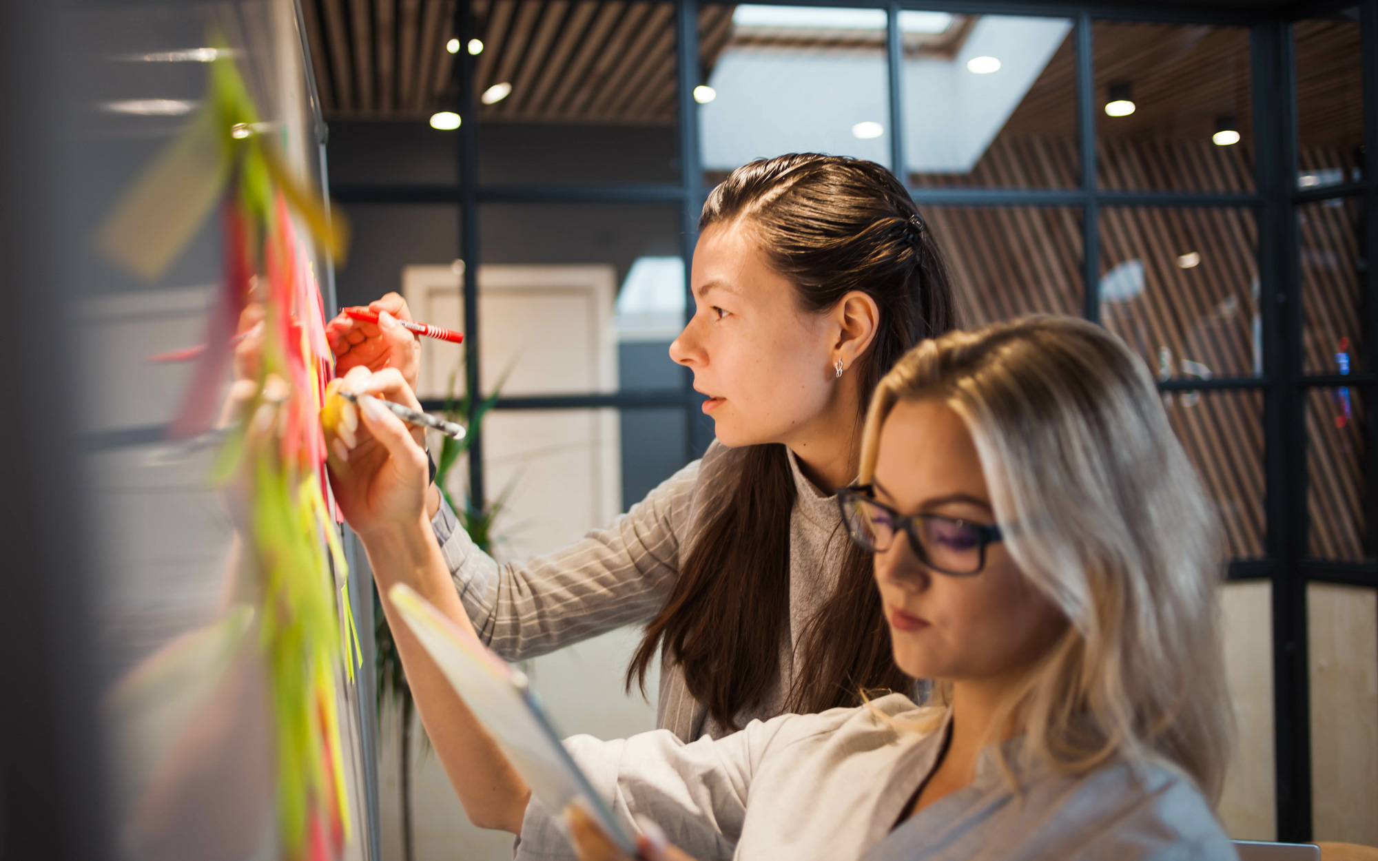 Zwei Frauen arbeiten konzentriert an einem Whiteboard mit bunten Haftnotizen, während sie Pläne und Ideen entwickeln.