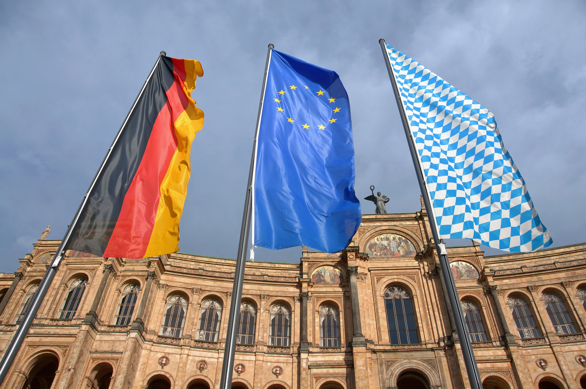 Drei Fahnen – die deutsche, die europäische und die bayerische – vor einem historischen Gebäude.