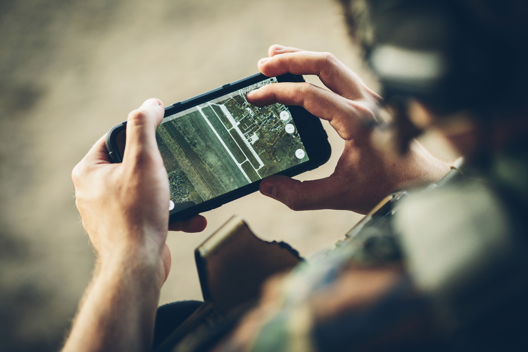 Eine Person betrachtet eine Satellitenkarte auf einem Smartphone, möglicherweise in einer taktischen oder militärischen Umgebung, basierend auf der Kleidung und dem Hintergrund.